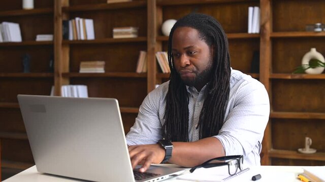Busy young African-American male office worker with dreadlocks removed glasses sitting at the desk and typing on laptop, student stressing in the library, taking an online exam, working on a project