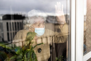 Senior woman with face mask looking out of window at home
