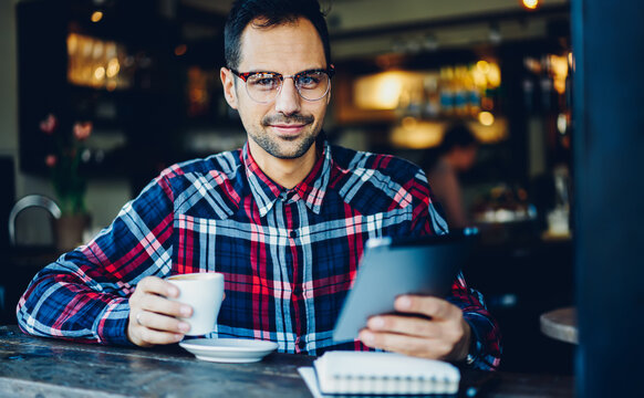 Portrait of handsome man looking at camera during coffee break for updating application on digital tablet, Caucasian male blogger in eyewear connecting to free 4G high speed internet on gadget
