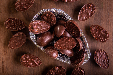Top view of a close up of mini stuffed chocolate easter eggs inside of a half of an easter egg wrapped in an aluminium paper. Some mini stuffed easter eggs around it on a wooden table.