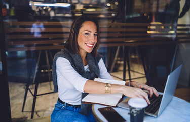 Smiling caucasian female typing on laptop computer enjoying distance job and wireless connection, cheerful businesswoman satisfied with online distance job sitting on cafe terrace look at camera