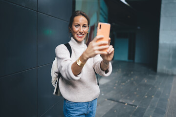 Half length portrait of cheerful female tourist smiling at camera while using smartphone gadget for...