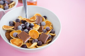 Pancake cereal in a plate with blueberries and chocolate on a pink table.Trendy modern breakfast with little pancakes.