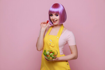 Woman with purple hair and bright makeup. Modern worker in fashionable work clothes showing product on pink background. Portrait of beautiful smiling female holding fresh vegetable salad bowl.