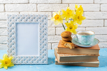 Spring flowers daffodils bouquet, white frame, coffee cup, books, cookies on blue table over white brick wall. Reading and breakfast, cozy home interior. Spring, hygge, spring holidays, read books