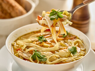 Soup with noodle, pieces of chicken and vegetables in white bowl served with spices and breads on light wooden background.