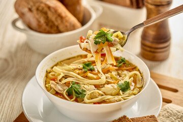 Soup with noodle, pieces of chicken and vegetables in white bowl served with spices and breads on light wooden background.