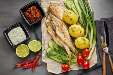 Fried smelts serving with potatoes, vegetables, lime and sauces. Grey slate background. Top view