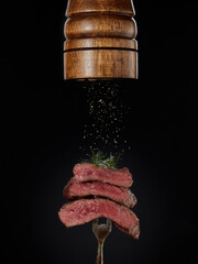Steak menu. Milled spices falling from pepper mill on grilled pieces of beef steak medium rare on fork on black background.