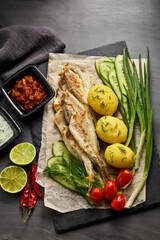 Fried smelts serving with potatoes, vegetables, lime and sauces. Grey slate background. Top view