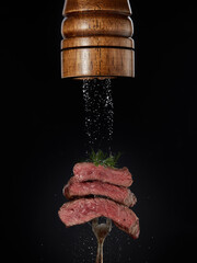 Steak menu. Milled salt falling from  mill on grilled pieces of beef steak medium rare on fork on black background.