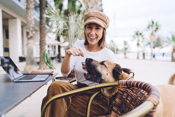 Portrait of cheerful caucasian female enjoying working with puppy in pet friendly cafe using laptop computer for freelancing, smiling woman 20s feeling happy sitting with cute dog on remote job