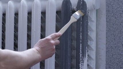male hand with a long brush with a curved end for painting in hard-to-reach places in the process of applying gray paint on a white home heating radiator, repainting a room in gray