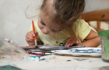 Little girl draws with paints sitting at the table in the room. Home education.