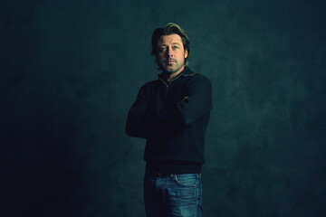 Middle aged man with stubble beard in green woolen sweater and jeans in front of grey wall.