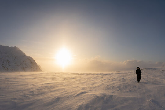 Scenic picture of large Sunset halo in winter mountains, selective focus because of strong snow drift wind. A person in black snowmobile overall and goggles walks against strong wind. Lapland Sweden