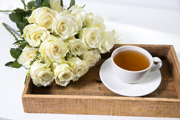 A bouquet of white roses in a vintage glass vase, a wooden tray with a cup of tea, a faience figurine of an angel. Greeting card for mother's day.