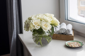 A bouquet of white roses in a round glass vase on the window with gray curtains. Tray with white small hand towels in the spa salon.