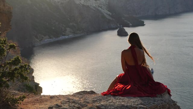 A young woman dressed in a red dress is sitting next to the sea on a rock, A girl is enjoying the view of the sea and the sunrise, her hair is blowing in the wind, the hem of the dress is developing.