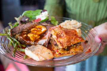 Clear glass plate of delicious food being held out with steak, chicken, and lasagna on it.