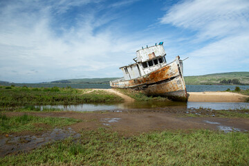 Abandoned fishing boat, shipwrecked and washed up on sandbar in muddy bog and grass. Blue sky with...