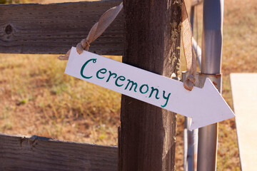 white painted arrow sign hanging on wood fence post says Ceremony