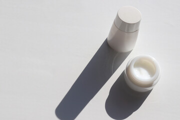 Face cream in glass jar and bottle of lotion on white table background. Hard light, shadow. Natural beauty products for skin and body care. Summer lifestyle. Top view, copy space
