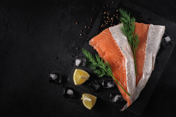 sliced fresh raw salmon lying on a stone cutting board with lemon wedges, dill, spices and ice cubes on a black textured concrete background. top view. beautiful sea food concept with copy space