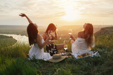 Three young happy girlfriends in elegant dresses is having picnic on the hill on sunset.