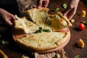 round four cheese pizza with basil sprig top view. soft focus on hands taking slices of pizza from a wooden plate on a brown background garnished with cherry tomatoes, parmesan cheese and basil sprigs