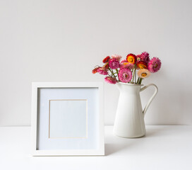 Close up of blank white picture frame on table with pink strawflowers in jug against wall (selective focus)
