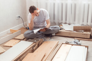 Bewildered man assembling new wooden furniture at home. Man reading instructions and misunderstand...