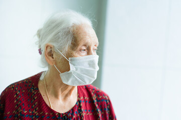 an elderly woman with gray hair 70-80 years old in a medical mask alone in the room stands at the window. Quarantine, coronavirus, stay at home, covid-19, risk group.