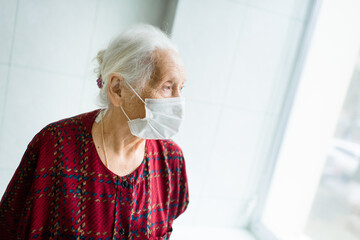 an elderly woman with gray hair 70-80 years old in a medical mask alone in the room stands at the window. Quarantine, coronavirus, stay at home, covid-19, risk group.