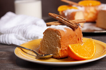 Concepto de un plato amarillo con un trozo de pastel con azúcar glash, ramas de canela, una rodaja de naranja y una cuchara de postre sobre un soporte de madera gris.