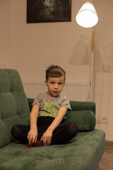 A boy in red socks is sitting cross-legged on a green sofa, and a floor lamp is lit next to him
