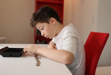 Boy with light skin and brown hair carefully examining coins in rows on a table. He is wearing a white shirt and sitting on a red chair.