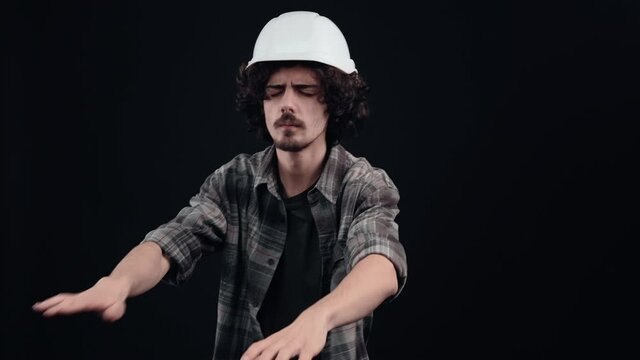 Puzzled hipster with outstretched hands looking for things in the dark. Young man with curly hair wears a special helmet on his head, dressed in a plaid shirt, on a black background, in the studio