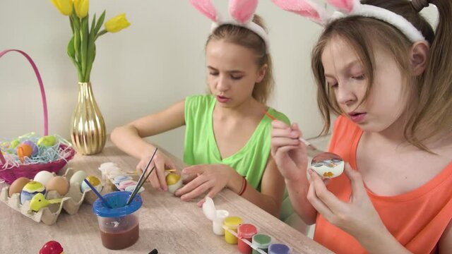 Girls decorate Easter eggs with paint. The girl opens the yellow paint. Easter holiday. On the table is a basket of Easter eggs and a vase of yellow tulips. 4k video.