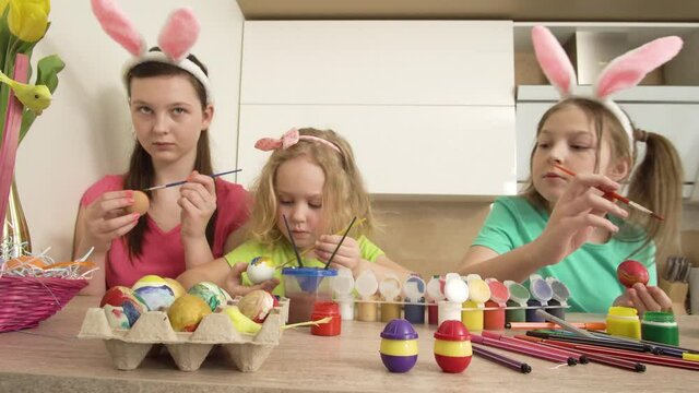 Three beautiful cute girls are decorating Easter eggs at home in the kitchen. 4k video.