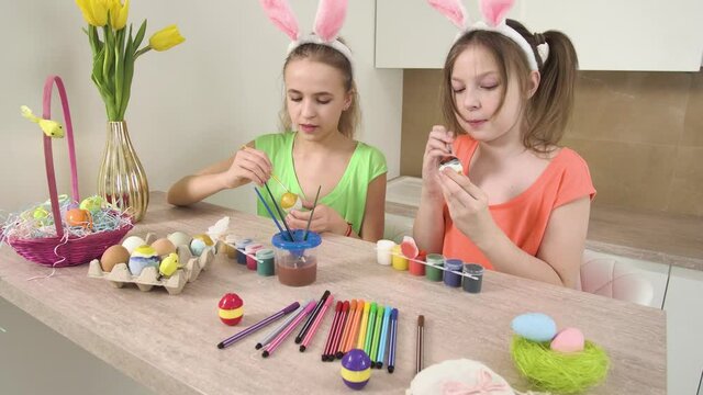 Girls with rabbit ears on their heads decorate Easter eggs with paint in the kitchen. Easter holiday. On the table is a basket of Easter eggs and a golden vase with yellow tulips. 4k video.
