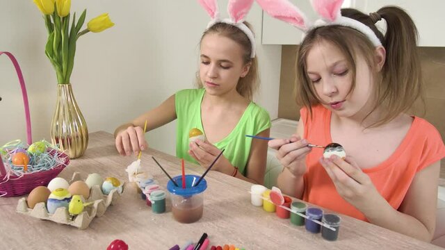 Two sisters decorate Easter eggs with colors and help each other. Easter holiday. On the table is a basket of Easter eggs and a vase of yellow tulips. 4k video.