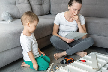 Cute adorable caucasian toddler boy kid sit on floor and help mom assembling furniture shelf with...