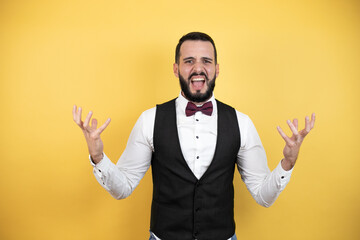 Young man with beard wearing bow tie and vest crazy and mad shouting and yelling with aggressive expression and arms raised. Frustration concept.