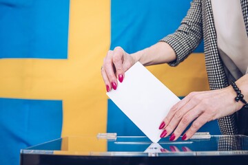 A womans hand puts her vote into the ballot box. Flag Sweden on background.
