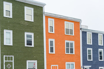 Street view of multiple colourful wooden buildings of various colours, green, orange and blue. The small structures have double hung windows and wooden doors. There's the grey sky in the background.