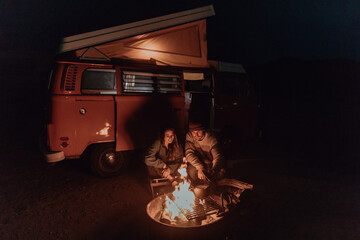 Young couple cooking camp fire by recreational vehicle at night, Jalama, California, USA