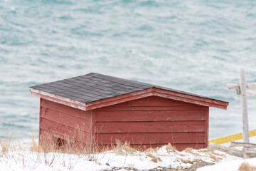 A small red wooden worn and weathered fishing shed or boathouse. The exterior has red boards and a small window with a black roof. There's a stormy and teal colour sea with snow near the building. 