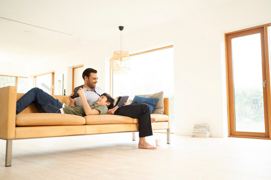 Boy and father using digital tablets whilst reclining on sofa