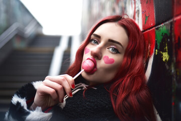 A glamorous teenage girl with red hair licks a sweet strawberry candy near a wall of graffiti.